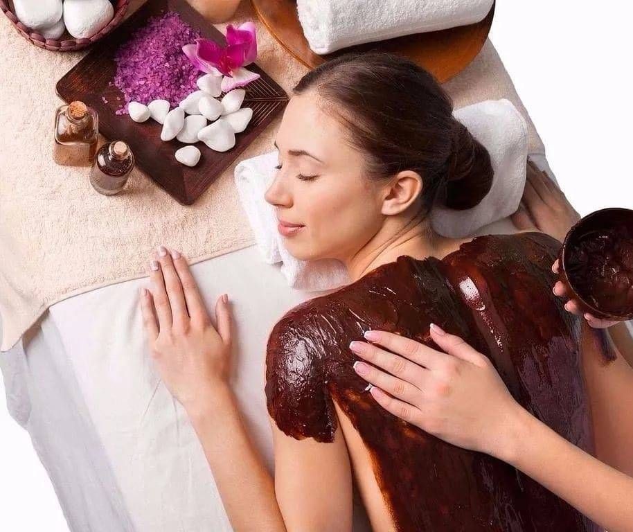 Woman receiving a relaxing back treatment with chocolate mask, surrounded by spa materials including oils and stones.